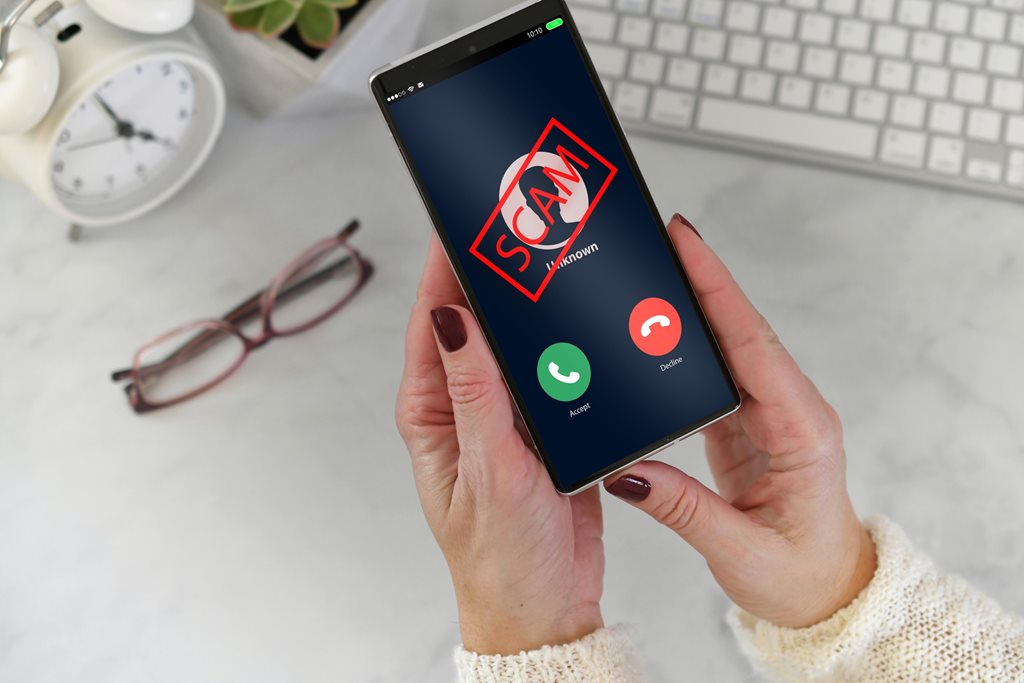 Hands holding a phone that shows a red scam alert over an incoming call with a background of a kitchen counter with a clock, glasses, and a keyboard sitting on it. 